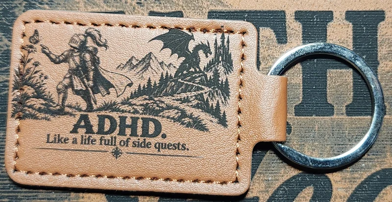 Photograph of a landscape tan stitched keychain showing an armored knight reaching toward a butterfly while a dragon and castle wait in the distance. Engraved text reads “ADHD. Like a life full of side quests.”