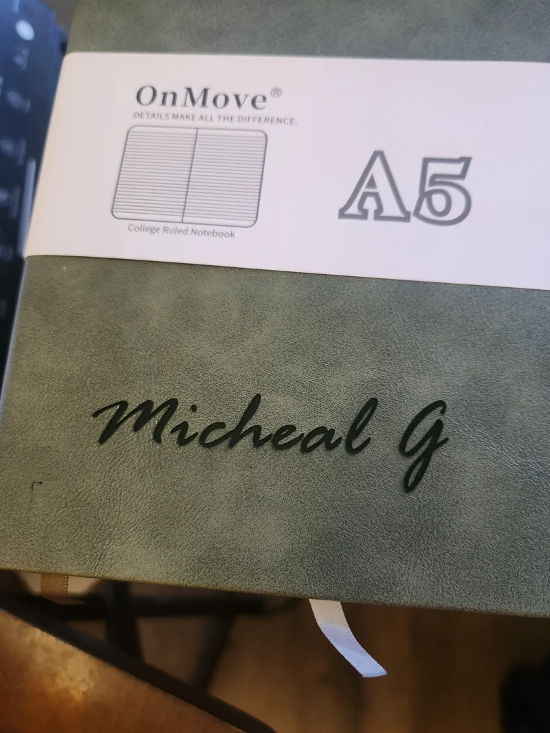 A close-up of a green notebook with a white label and the name 'Micheal G' embossed on it.