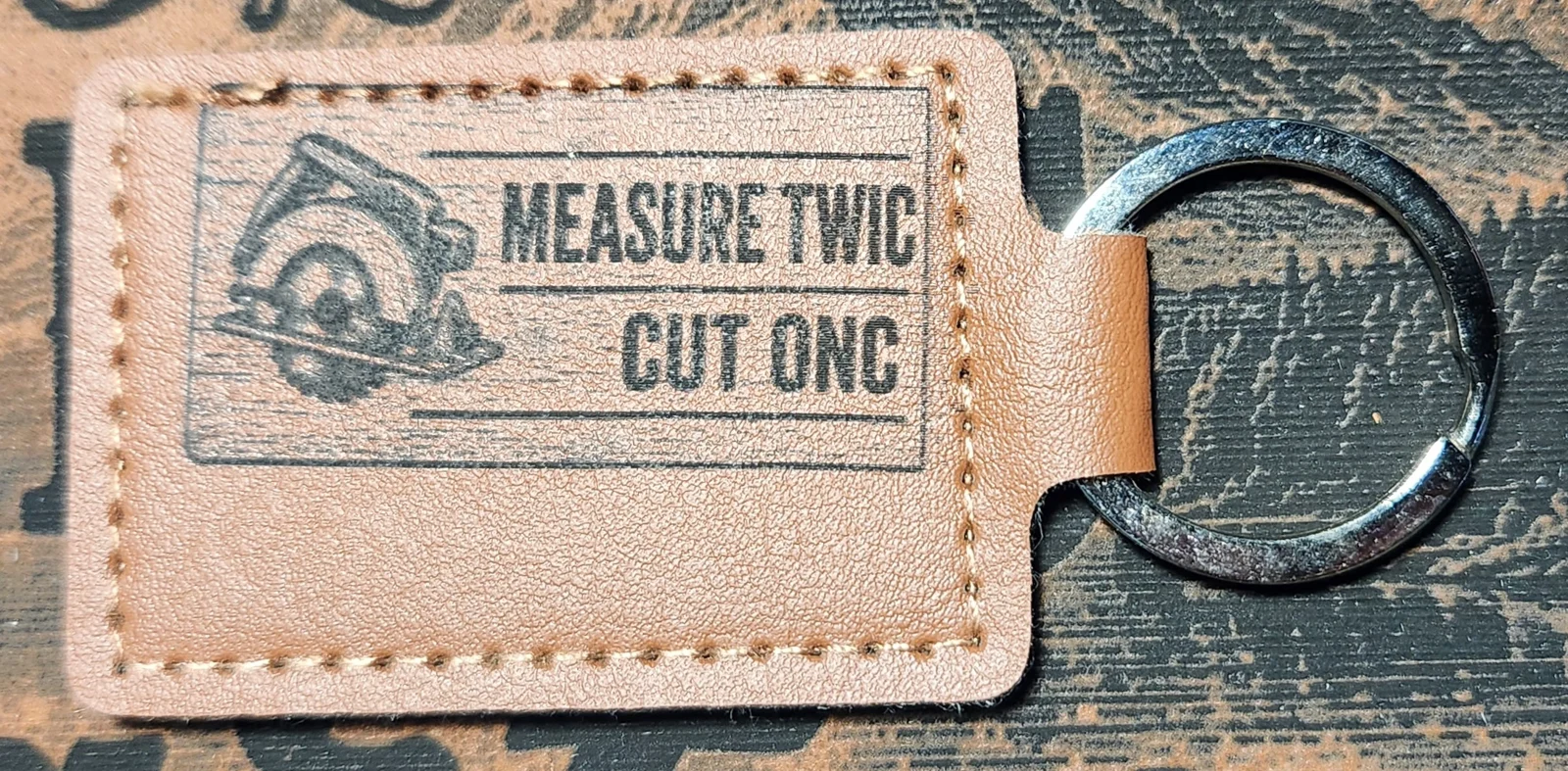 Photograph of a landscape tan stitched keychain with a circular saw and two lines of abbreviated lettering. Engraved text reads “MEASURE TWIC CUT ONC.”