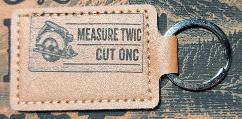 Photograph of a landscape tan stitched keychain with a circular saw and two lines of abbreviated lettering. Engraved text reads “MEASURE TWIC CUT ONC.”