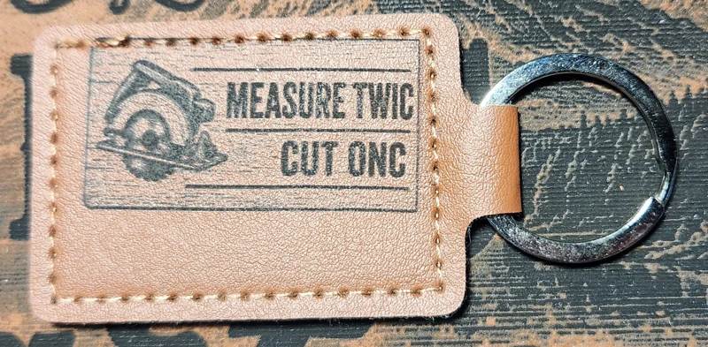 Photograph of a landscape tan stitched keychain with a circular saw and two lines of abbreviated lettering. Engraved text reads “MEASURE TWIC CUT ONC.”