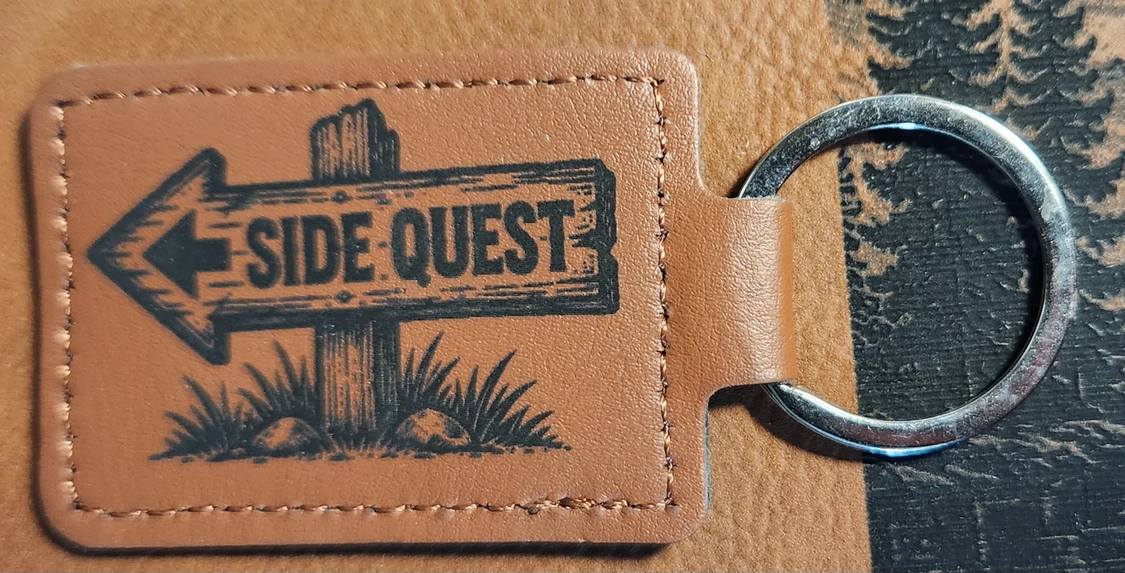 Photograph of a landscape tan stitched keychain with a rustic wooden sign pointing left and grass around its post. Engraved text reads “SIDE QUEST.”