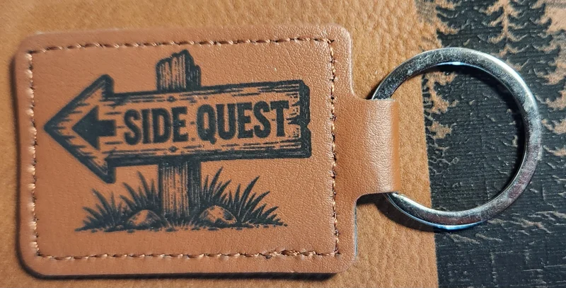 Photograph of a landscape tan stitched keychain with a rustic wooden sign pointing left and grass around its post. Engraved text reads “SIDE QUEST.”