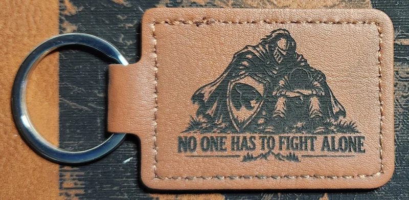 Tan stitched landscape keychain engraved with two armored figures beside a shield and the words NO ONE HAS TO FIGHT ALONE.