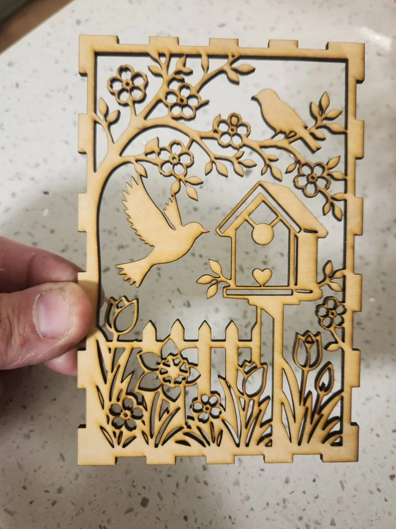 A laser-cut wooden panel with a design of a birdhouse, a flying bird, a perched bird, and floral motifs.