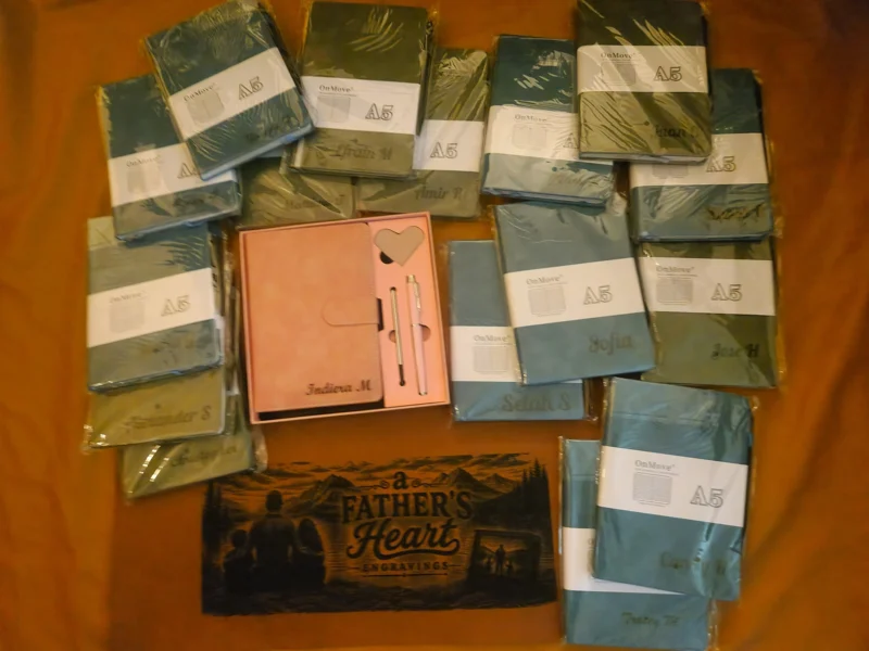 A collection of A5 notebooks in various shades of blue, green, and grey, neatly arranged on a wooden surface. A pink notebook with a heart-shaped cutout and two pens is placed in the center. Below the notebooks, there is a book titled 'A Father's Heart Engravings' with an illustration of two figures sitting on a bench.