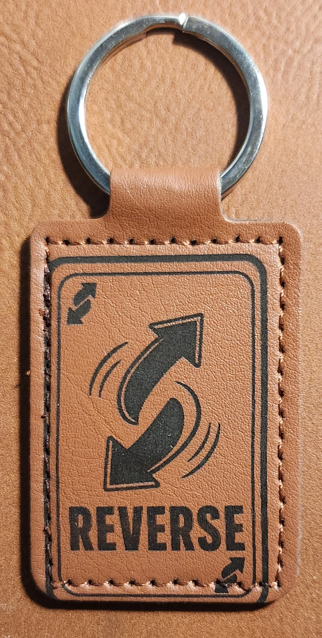 Tan stitched portrait keychain engraved with two curved arrows, a playing-card border, and the word REVERSE.
