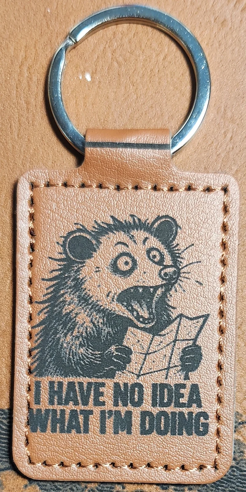 Photograph of a portrait tan stitched keychain with a wide-eyed open-mouthed cartoon animal holding an unfolded map above the words. Engraved text reads “I HAVE NO IDEA WHAT I’M DOING.”