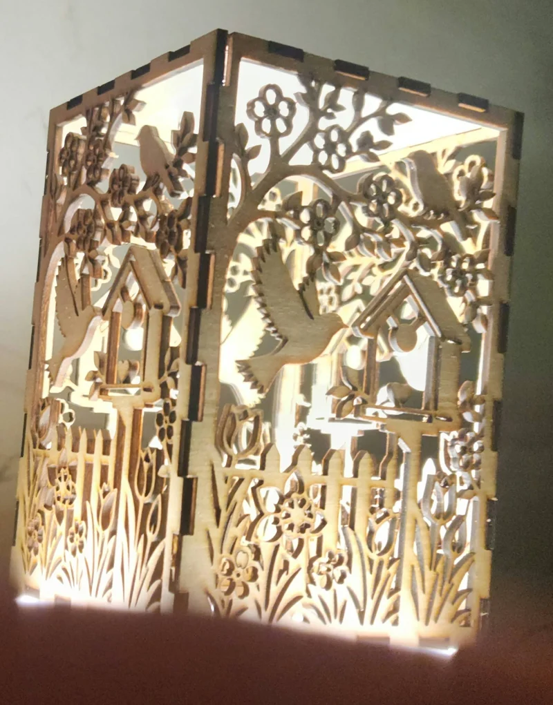 A laser-cut wooden artwork with layered panels featuring birds, flowers, and birdhouses, backlit to create a glowing effect.