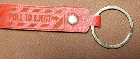 Photograph of a long narrow red strap keychain with an arrow, striped border, metal rivet, and split ring. Engraved text reads “PULL TO EJECT →.”