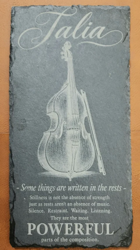 Close-up of a personalized engraved black slate for Talia featuring a double bass and bow with a message about the power of stillness, listening and rests in music.