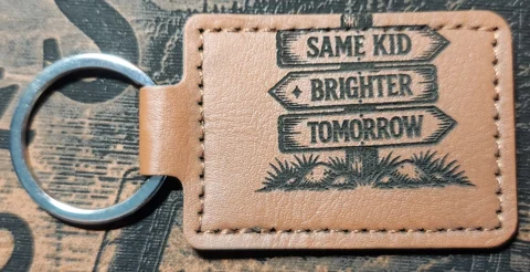 Tan stitched landscape keychain showing a rustic three-arrow signpost with the words SAME KID, BRIGHTER, and TOMORROW above grass and stones.