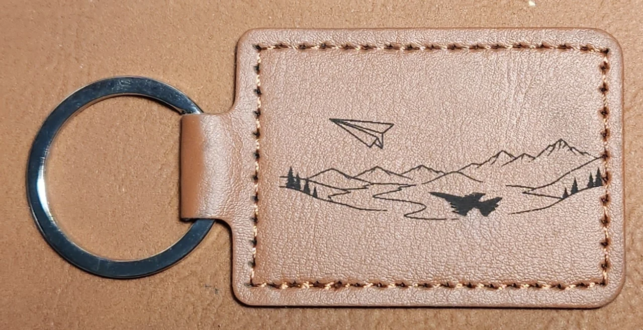 Tan stitched landscape keychain showing a paper airplane above mountain outlines, scattered pine trees, and a jet-shaped silhouette below.
