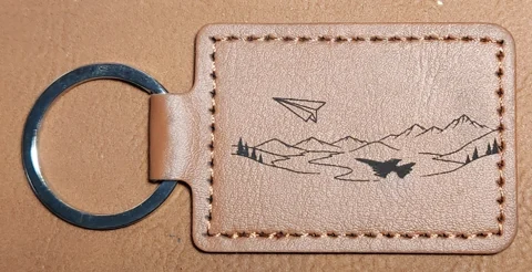 Tan stitched landscape keychain showing a paper airplane above mountain outlines, scattered pine trees, and a jet-shaped silhouette below.