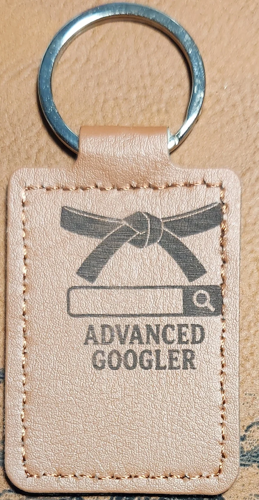 Photograph of a portrait tan stitched keychain featuring a tied martial-arts-style belt above a search bar and two lines of text. Engraved text reads “ADVANCED GOOGLER.”