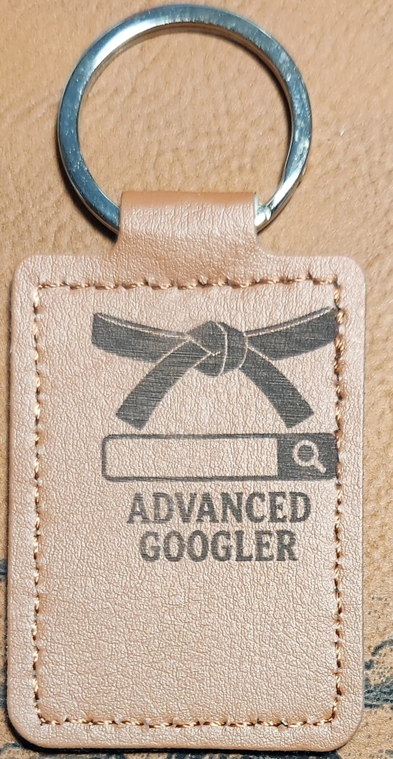 Photograph of a portrait tan stitched keychain featuring a tied martial-arts-style belt above a search bar and two lines of text. Engraved text reads “ADVANCED GOOGLER.”