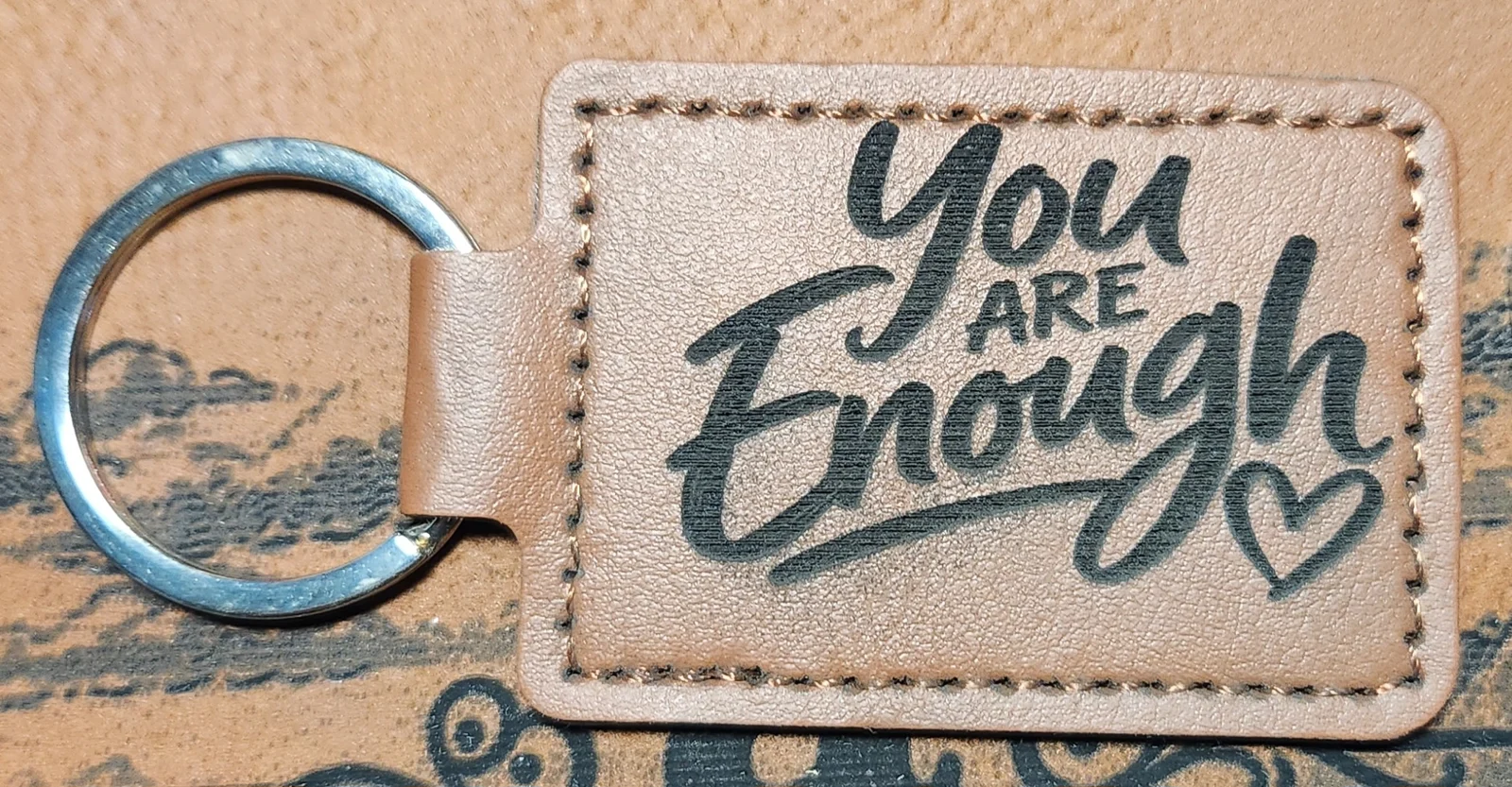 Tan stitched landscape keychain engraved with the words You ARE Enough in mixed script and block lettering beside an outlined heart.