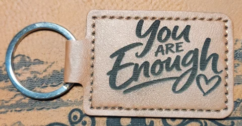Tan stitched landscape keychain engraved with the words You ARE Enough in mixed script and block lettering beside an outlined heart.