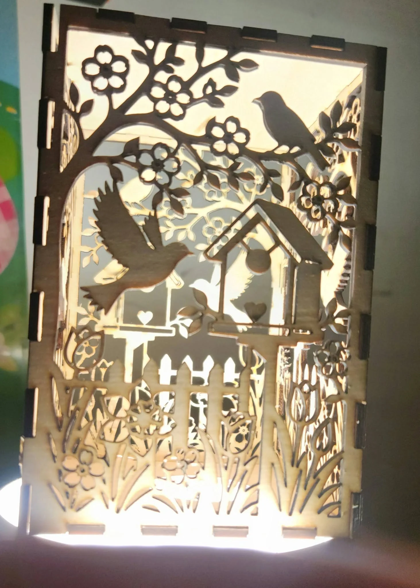 A laser-cut wooden panel with layered silhouettes of birds, flowers, and a birdhouse, backlit to create a glowing effect.
