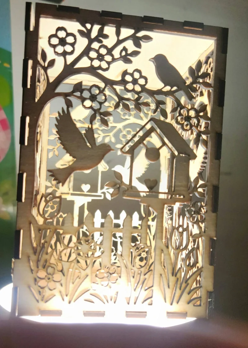 A laser-cut wooden panel with layered silhouettes of birds, flowers, and a birdhouse, backlit to create a glowing effect.