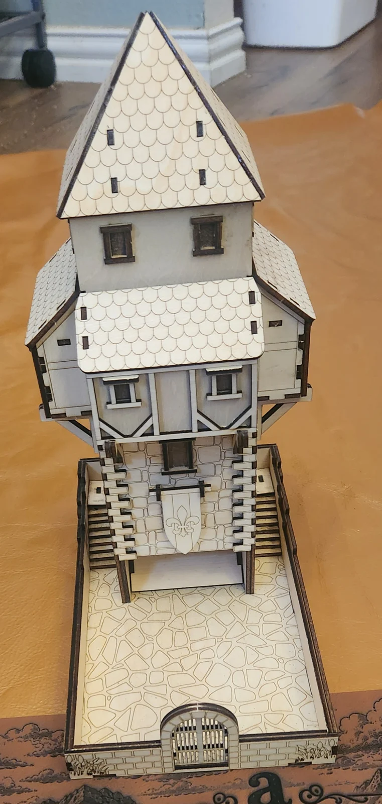 A detailed model of a medieval-style house with a steep, pointed roof and a stone base featuring a fleur-de-lis emblem.