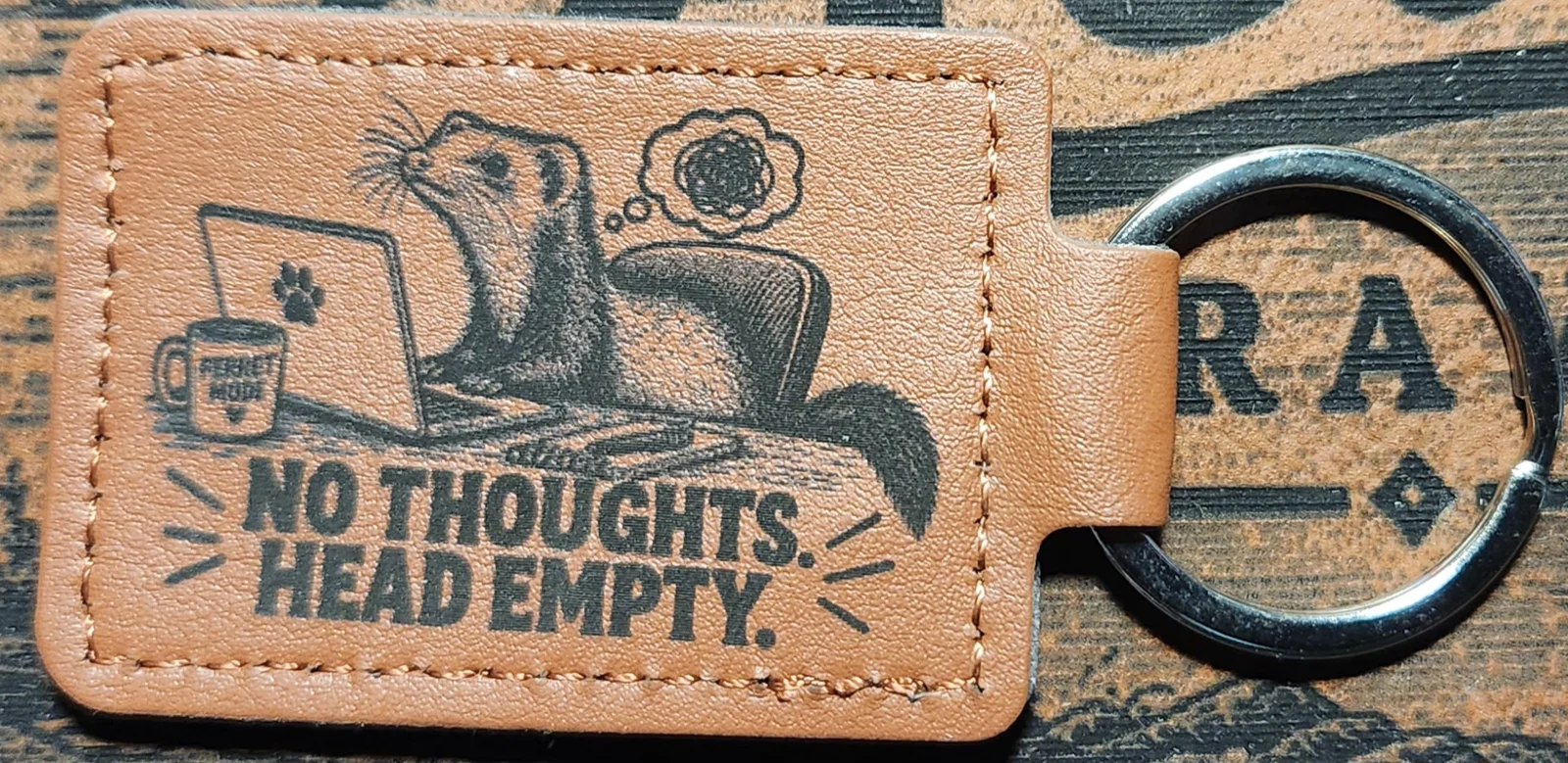 Photograph of a landscape tan stitched keychain showing a long-bodied ferret seated at a laptop with a mug and a scribbled thought bubble. Engraved text reads “NO THOUGHTS. HEAD EMPTY.”