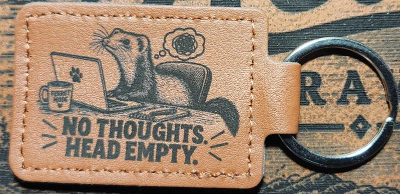 Photograph of a landscape tan stitched keychain showing a long-bodied ferret seated at a laptop with a mug and a scribbled thought bubble. Engraved text reads “NO THOUGHTS. HEAD EMPTY.”