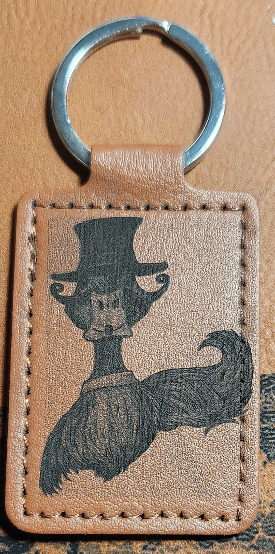 Photograph of a portrait tan stitched keychain featuring a front-facing duck with a tall top hat and an oversized curled mustache.