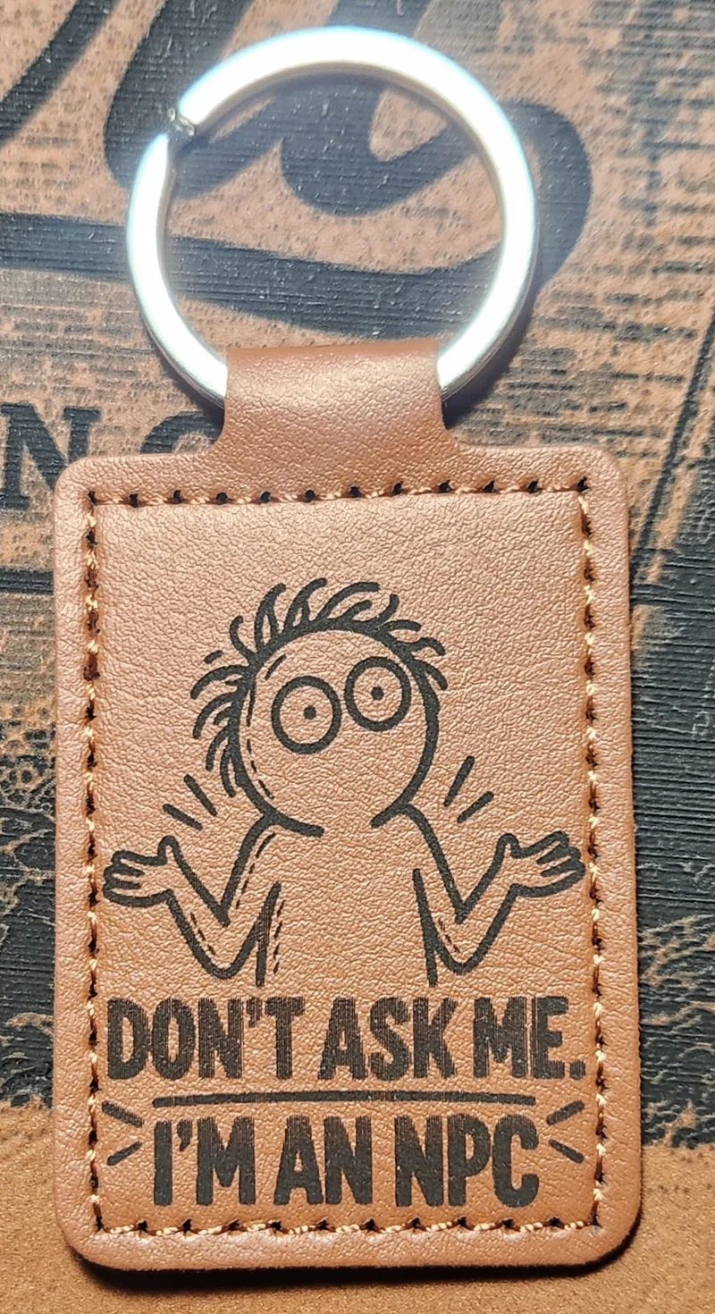 Photograph of a portrait tan stitched keychain with a wide-eyed cartoon person shrugging above the words. Engraved text reads “DON’T ASK ME. I’M AN NPC.”
