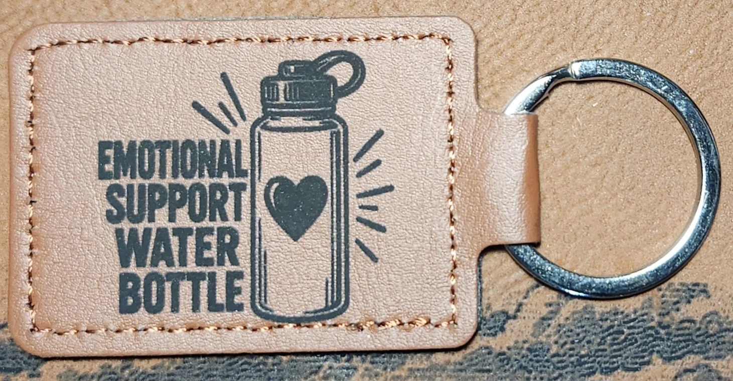 Photograph of a landscape tan stitched keychain with stacked text and a water bottle containing a heart. Engraved text reads “EMOTIONAL SUPPORT WATER BOTTLE.”