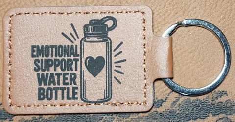 Photograph of a landscape tan stitched keychain with stacked text and a water bottle containing a heart. Engraved text reads “EMOTIONAL SUPPORT WATER BOTTLE.”