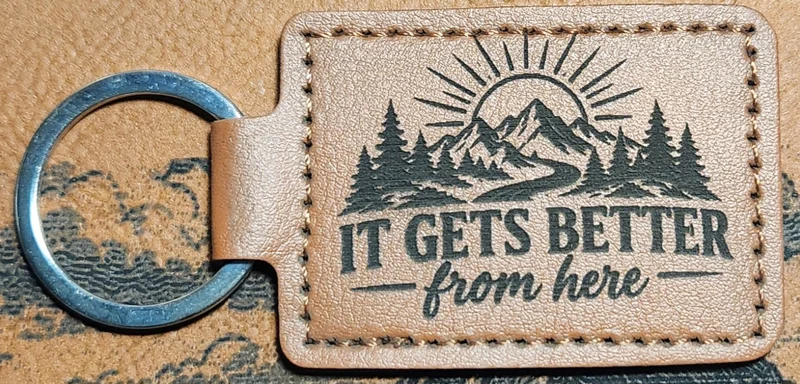 Tan stitched landscape keychain with mountains, pine trees, a winding path, sun rays, and the words IT GETS BETTER from here.