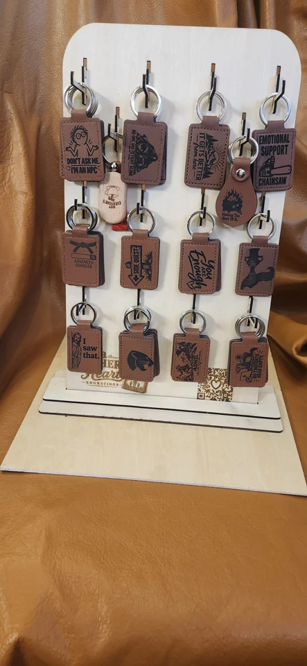 Freestanding wooden keychain display with rows of hooks holding engraved keychains featuring humorous and encouraging designs. A Father’s Heart Engravings branding and a QR code appear near the base.