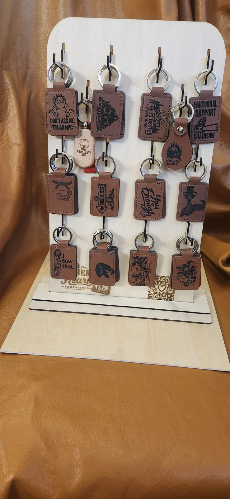 Freestanding wooden keychain display with rows of hooks holding engraved keychains featuring humorous and encouraging designs. A Father’s Heart Engravings branding and a QR code appear near the base.