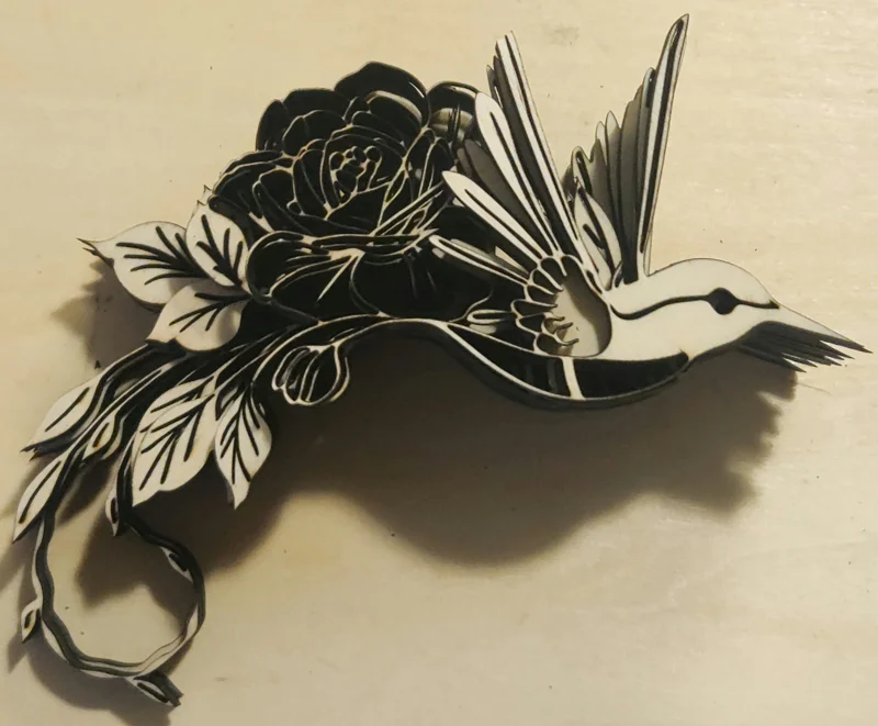 Layered laser-cut wooden artwork of a hummingbird flying beside a large rose with detailed leaves and flowers, created in memory of the artist's late mother and her love of hummingbirds