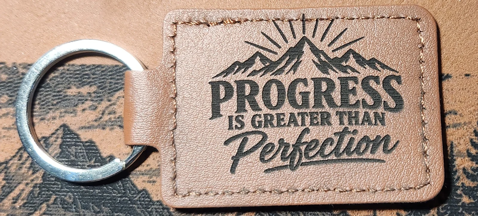 Tan stitched landscape keychain engraved with mountain peaks, sun rays, and the words PROGRESS IS GREATER THAN Perfection.