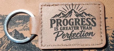 Tan stitched landscape keychain engraved with mountain peaks, sun rays, and the words PROGRESS IS GREATER THAN Perfection.
